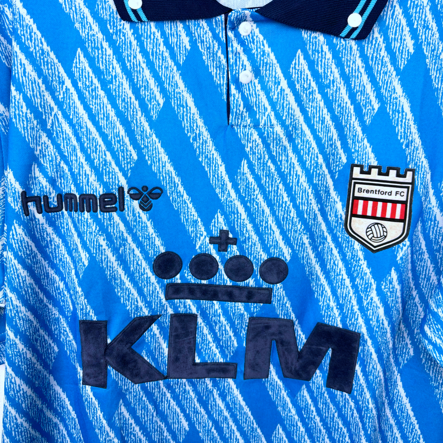 Close up of retro Brentford 1992-1993 away shirt. Geometrical sky-blue pattern and navy collar. Visible branding of Hummel, KLM, and Brentford FC crest.
