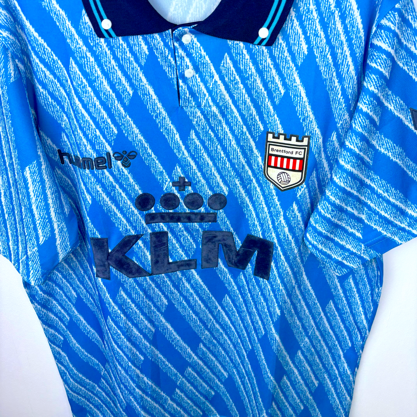 Downward view of Brentford 1992 away classic football shirt produced by Hummel. Geometrical sky-blue pattern and navy collar. KLM airlines sponsor.
