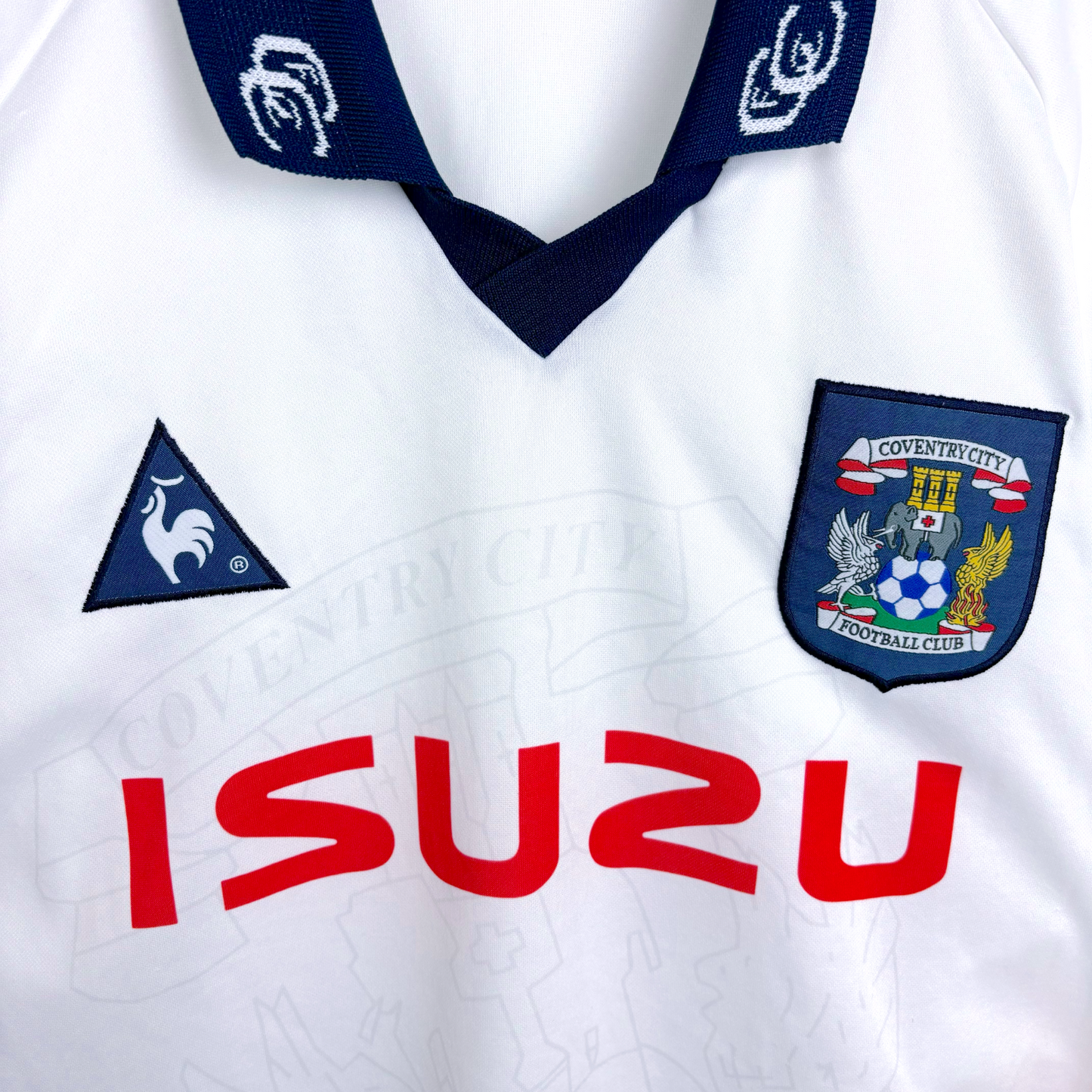 Close up of white Coventry City classic football shirt produced by le coq sportif. Embroidered Coventry and le coq sportif badges with red Izuzu sponsor.