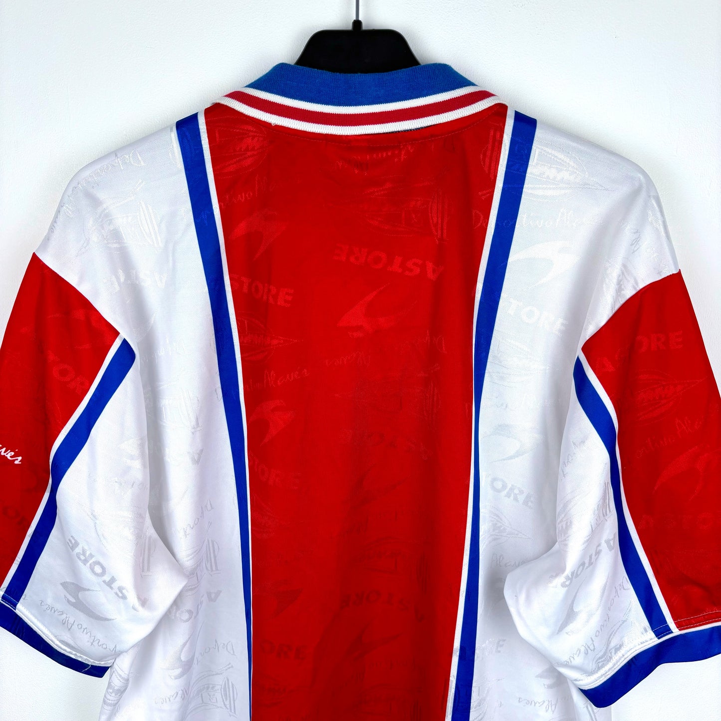 The image shows the back of a vintage Deportivo Alavés 1994-1999 Away Football Shirt by Astore, featuring red, white, and blue panels with a bold red center stripe, hanging on a black hanger against a white wall.