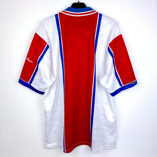 A vintage Deportivo Alavés 1994-1999 away shirt by Astore, featuring a red central panel, white sleeves and sides, blue vertical stripes, and a white collar with blue and red trim, displayed on a hanger.
