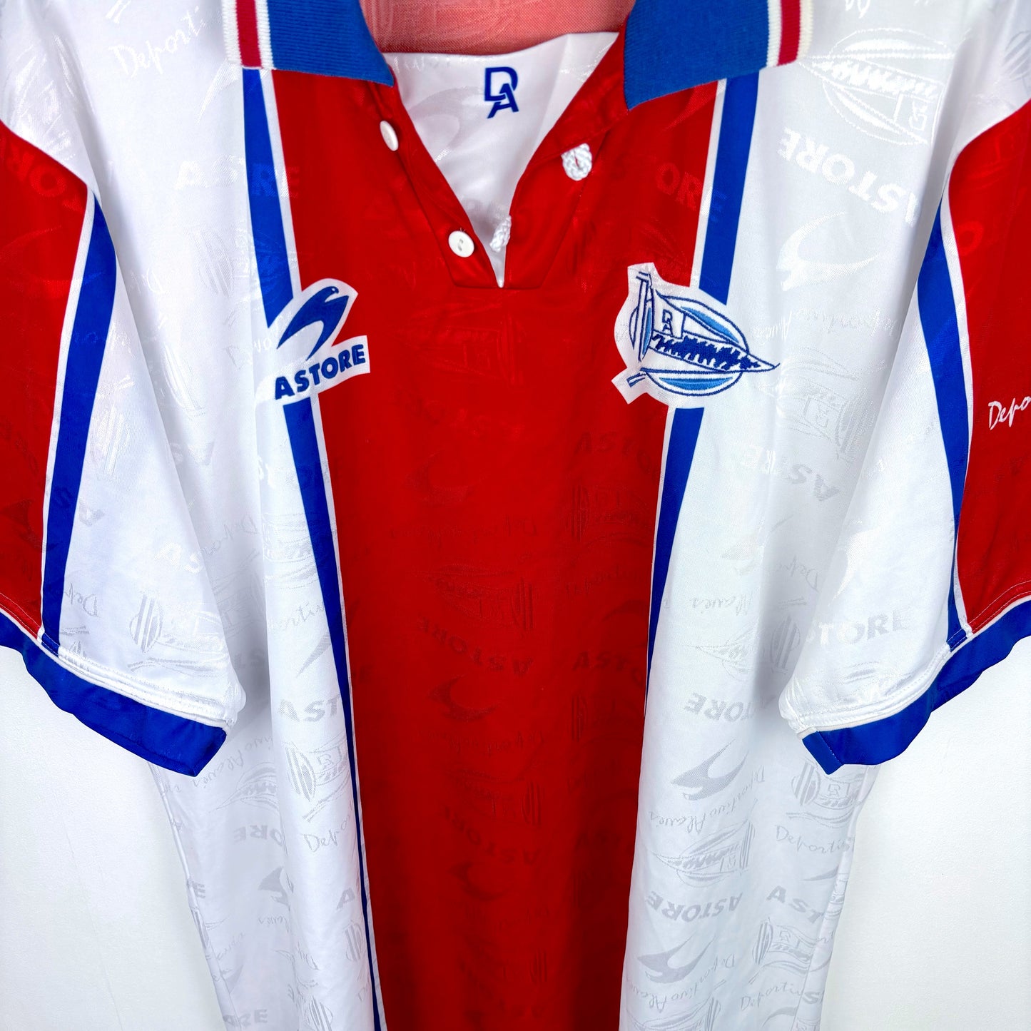 The Deportivo Alavés 1994-1999 Away Football Shirt by Astore features red, white, and blue vertical panels, blue collar and sleeve trim, and a stylized ship crest, replicating the classic away kit worn in Spain’s top flight.