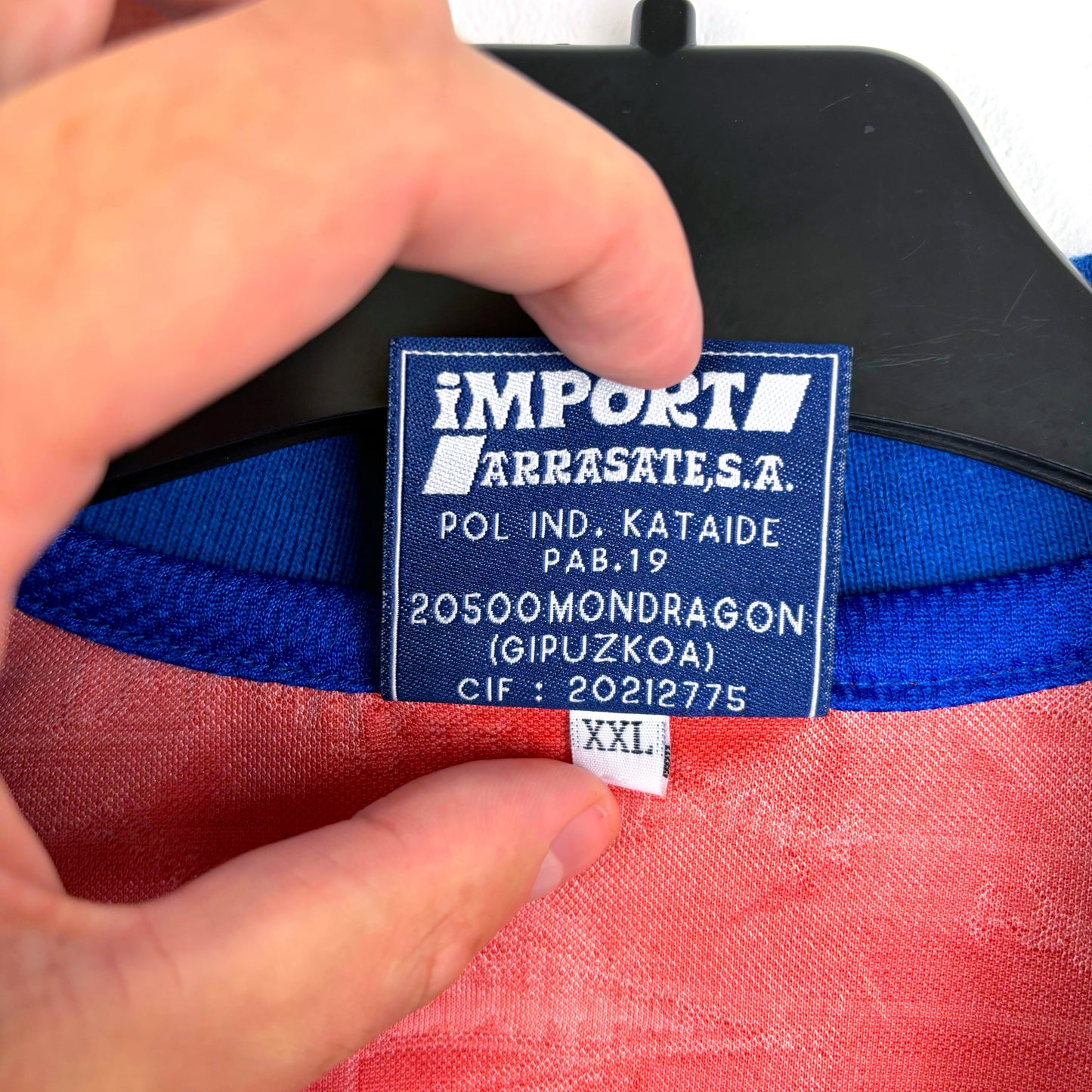 A hand holds a red Deportivo Alavés 1994-1999 away football shirt by Astore on a hanger, showing a blue and white label with company details and size XXL, from Spain's top flight.
