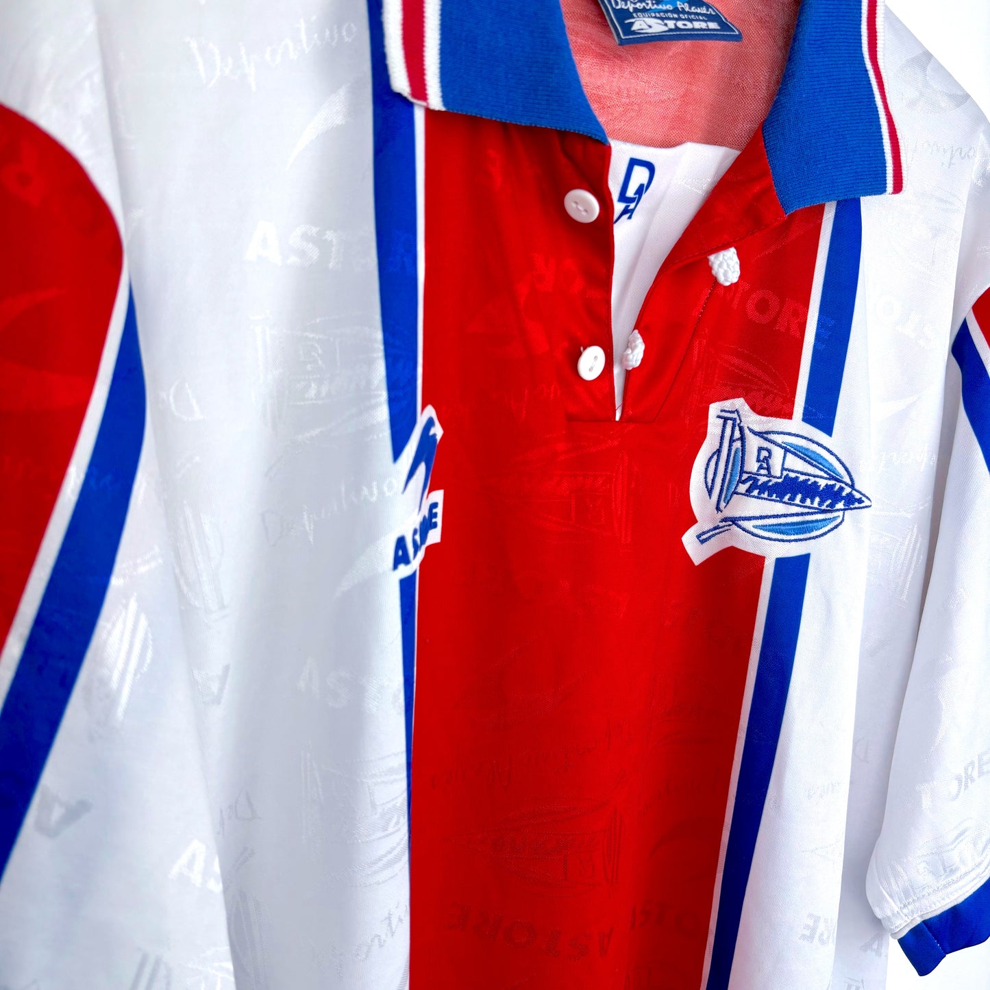 A close-up of a vintage Deportivo Alavés 1994-1999 away shirt by Astore, featuring red, white, and blue vertical stripes, blue collar, button placket, and a patch with a ship and "AS Deportivo" from Spain’s top flight.