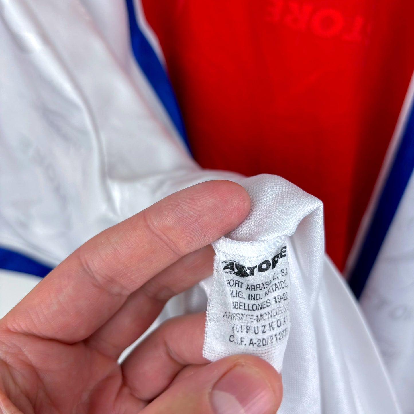 A hand holds a white tag attached to a shiny white Deportivo Alavés 1994-1999 Away Football Shirt by Astore. The shirt features blue trim, and the tag shows black printed Astore brand details, set against a red background.