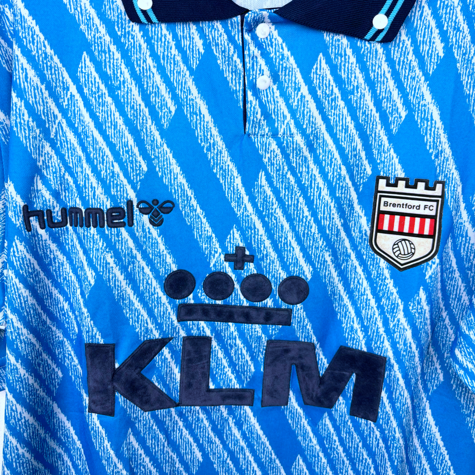 Close up of retro Brentford 1992-1993 away shirt. Geometrical sky-blue pattern and navy collar. Visible branding of Hummel, KLM, and Brentford FC crest.