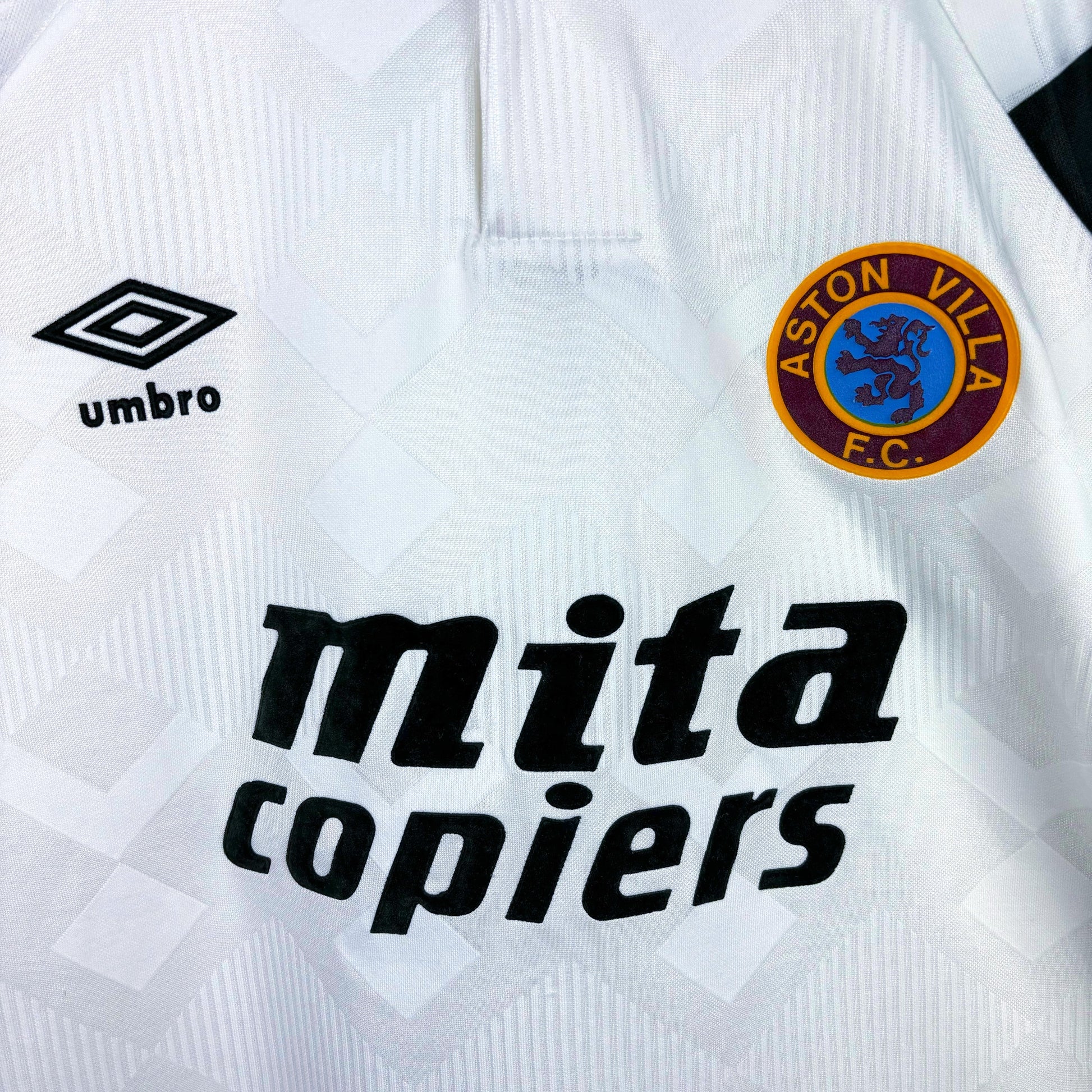 Close up of Aston Villa 1990-1993 away vintage football shirt made by Umbro. Sublimated jacquard pattern ion white body. Black and blue trim with black Mita Copiers sponsor and club crest to right.