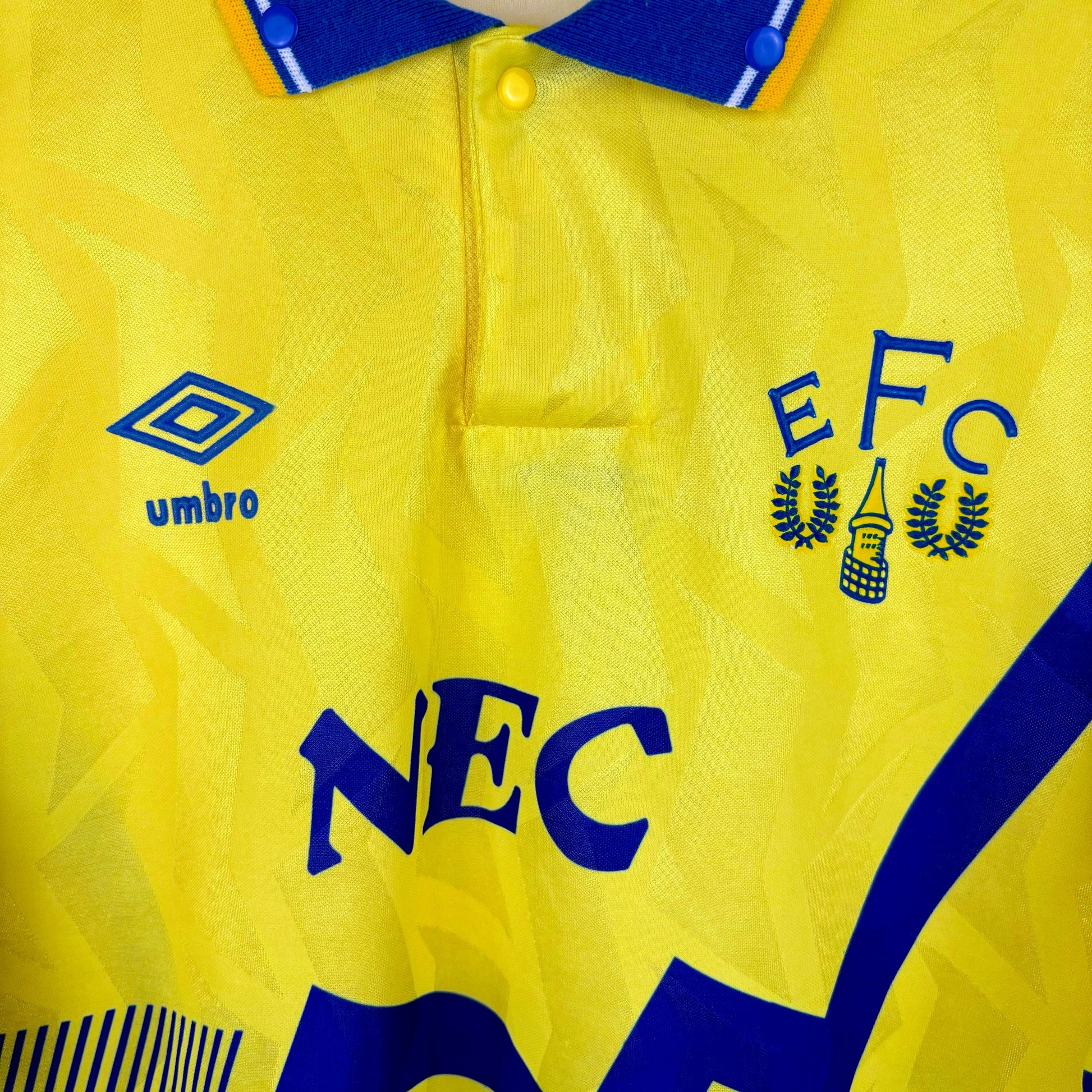 Close up view of Everton 1990-1992 away classic football shirt made by Umbro. Blue sublimated zig-zag across yellow body . NEC sponsor and old club crest.