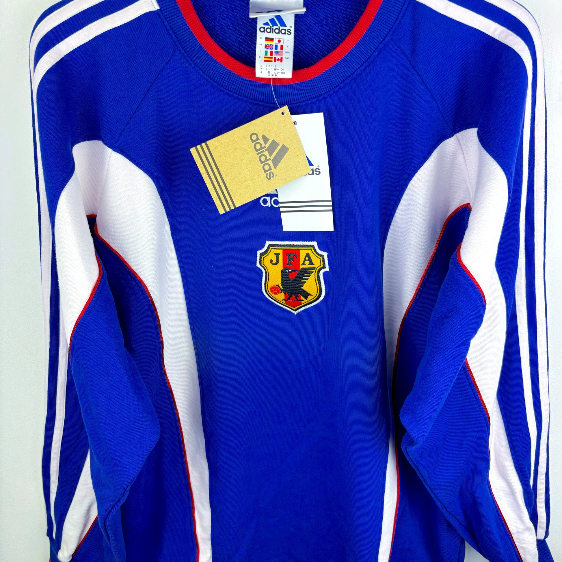 Side view of Japan 1999-2000 adidas retro soccer sweatshirt. Blue body with white vertical swooshes and red trim. Brand new with tags attached. 