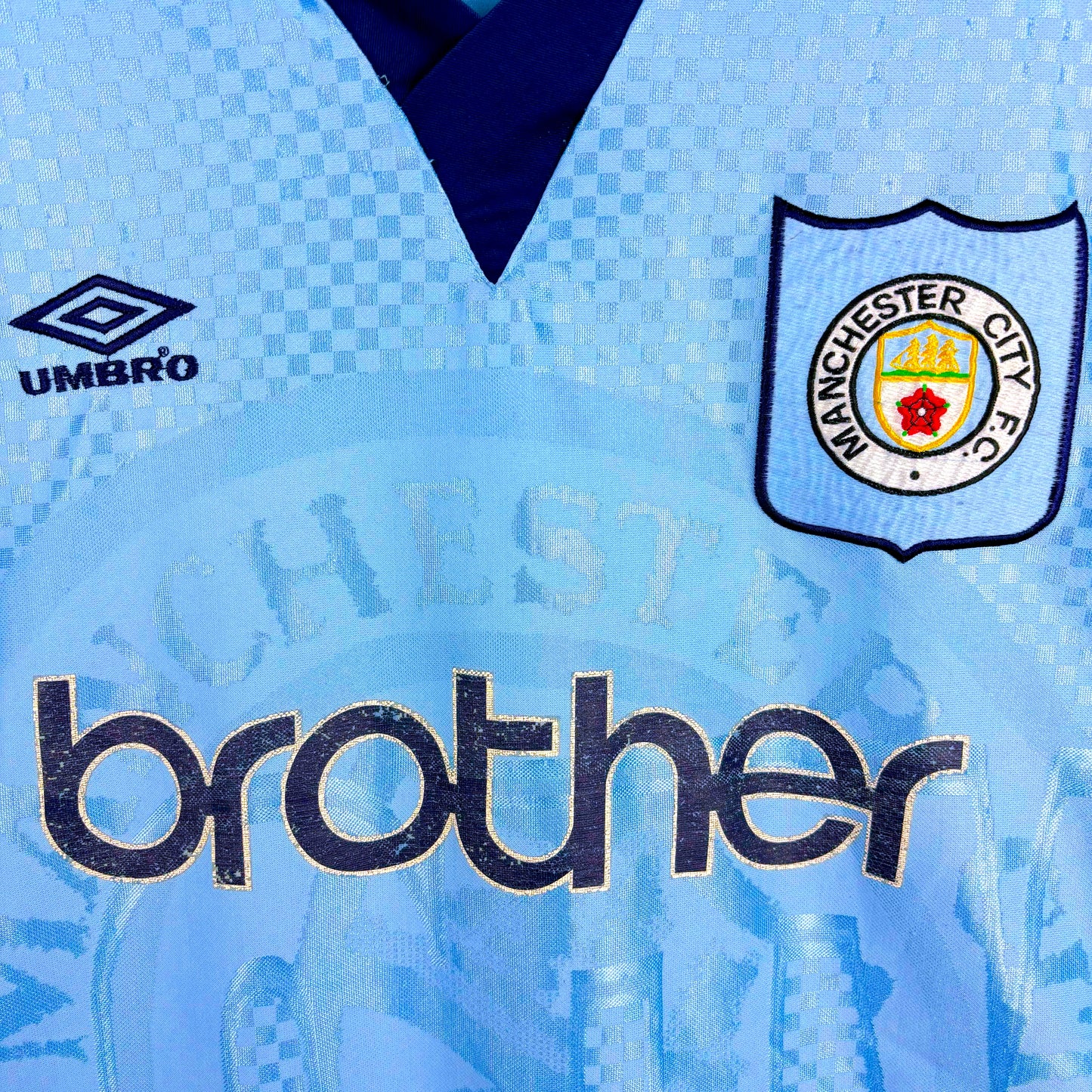 Close up of badges and Brother sponsor on Man City 1995-1997 Umbro home vintage football jersey. Sky blue body with sublimated checkered pattern.