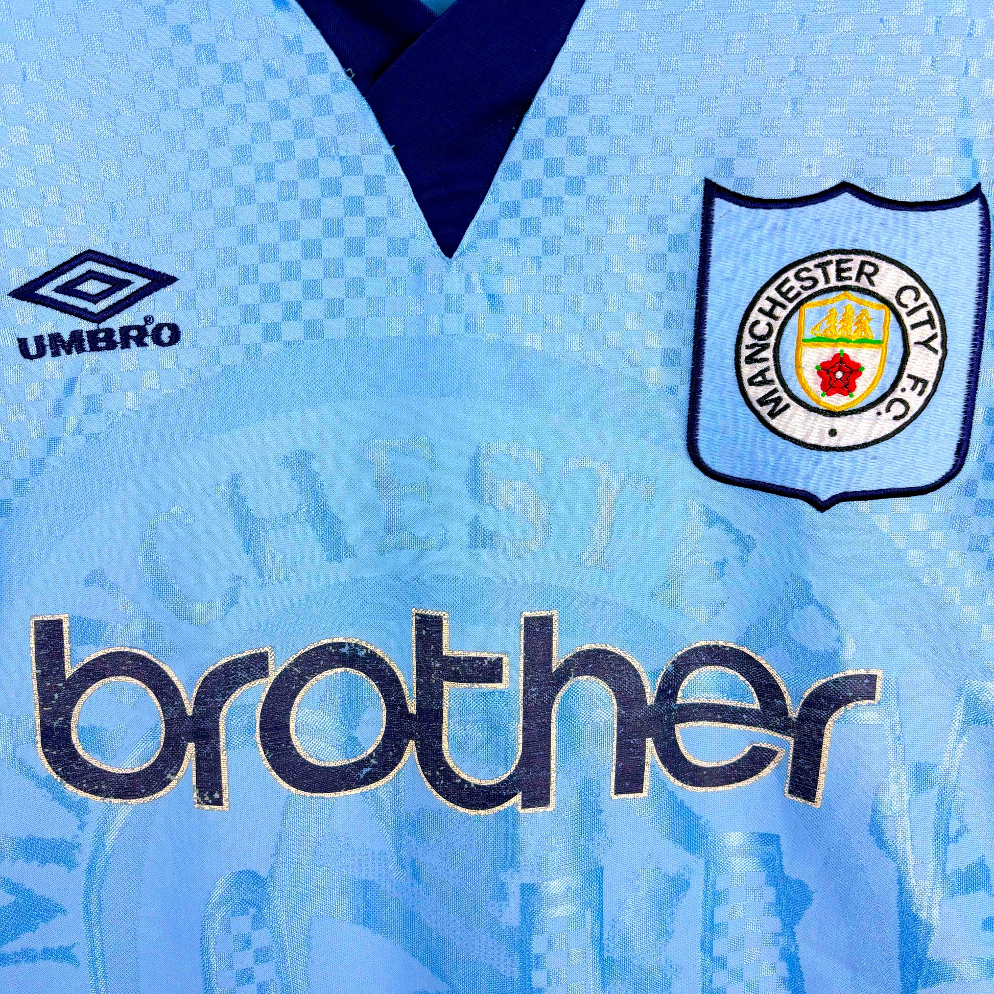 Close up of badges and Brother sponsor on Man City 1995-1997 Umbro home vintage football jersey. Sky blue body with sublimated checkered pattern.