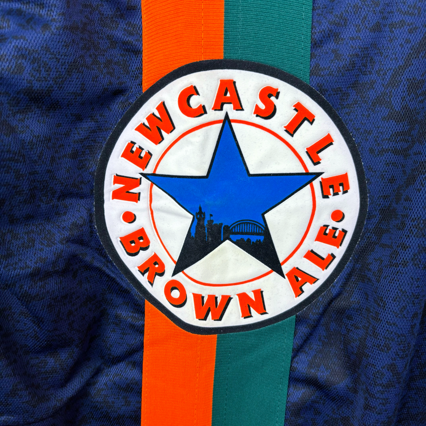 Sponsor close up of Newcastle United retro soccer jersey made by Adidas. Navy body featuring duel green and orange stripes and crew neck collar.