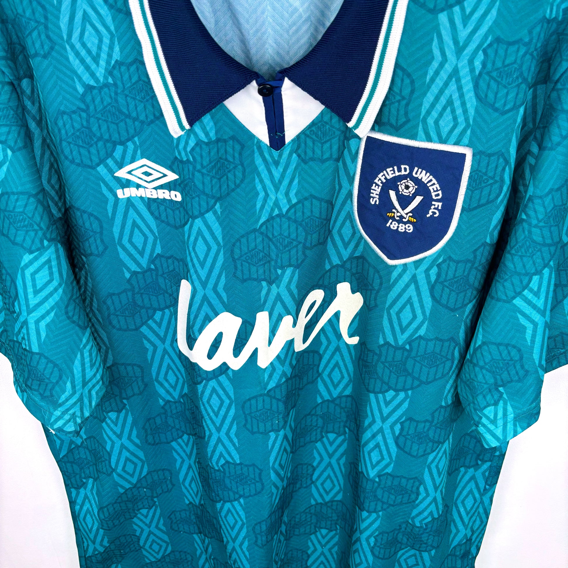 Downward view of Sheffield United 1994-1995 away retro football shirt made by Umbro. Sublimated Umbro pattern in teal/emerald with navy collar. Laver sponsor across chest.