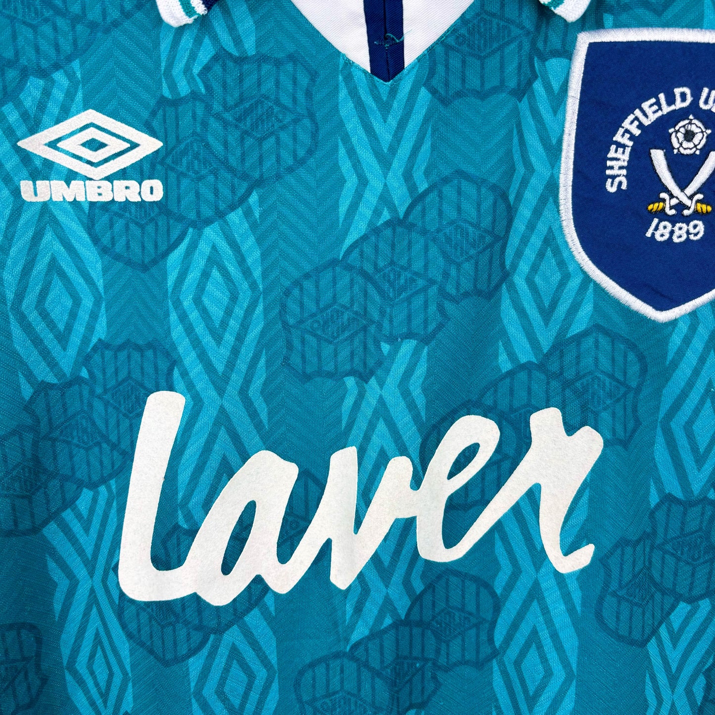Close up of Sheffield United 1994-1995 away vintage football shirt made by Umbro. Sublimated Umbro pattern in teal/emerald with navy collar. Laver and Umbro sponsors in white with navy embroidered club crest